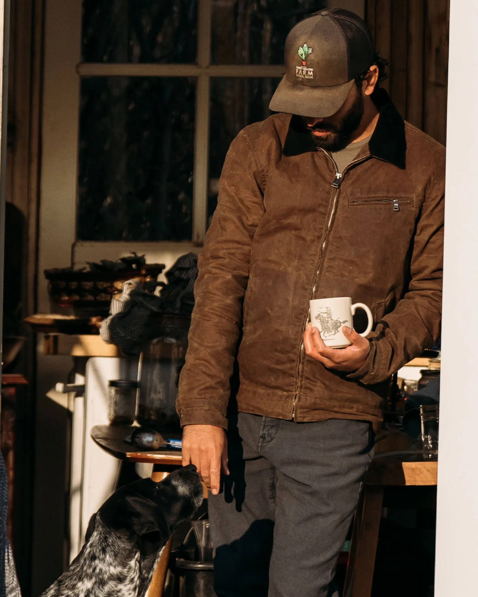 Man in brown jacket and gray cap holding a white mug and touching a black-and-white dog indoors.