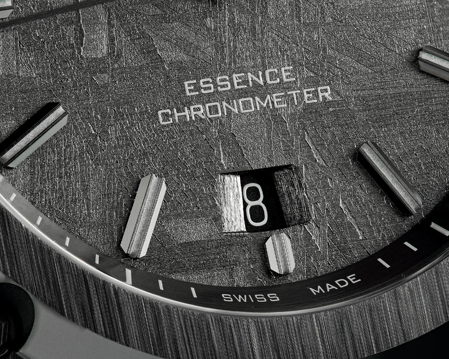 Close-up of a textured black watch dial with silver hour markers, date window showing "8," and text "ESSENCE CHRONOMETER" and "SWISS MADE.
