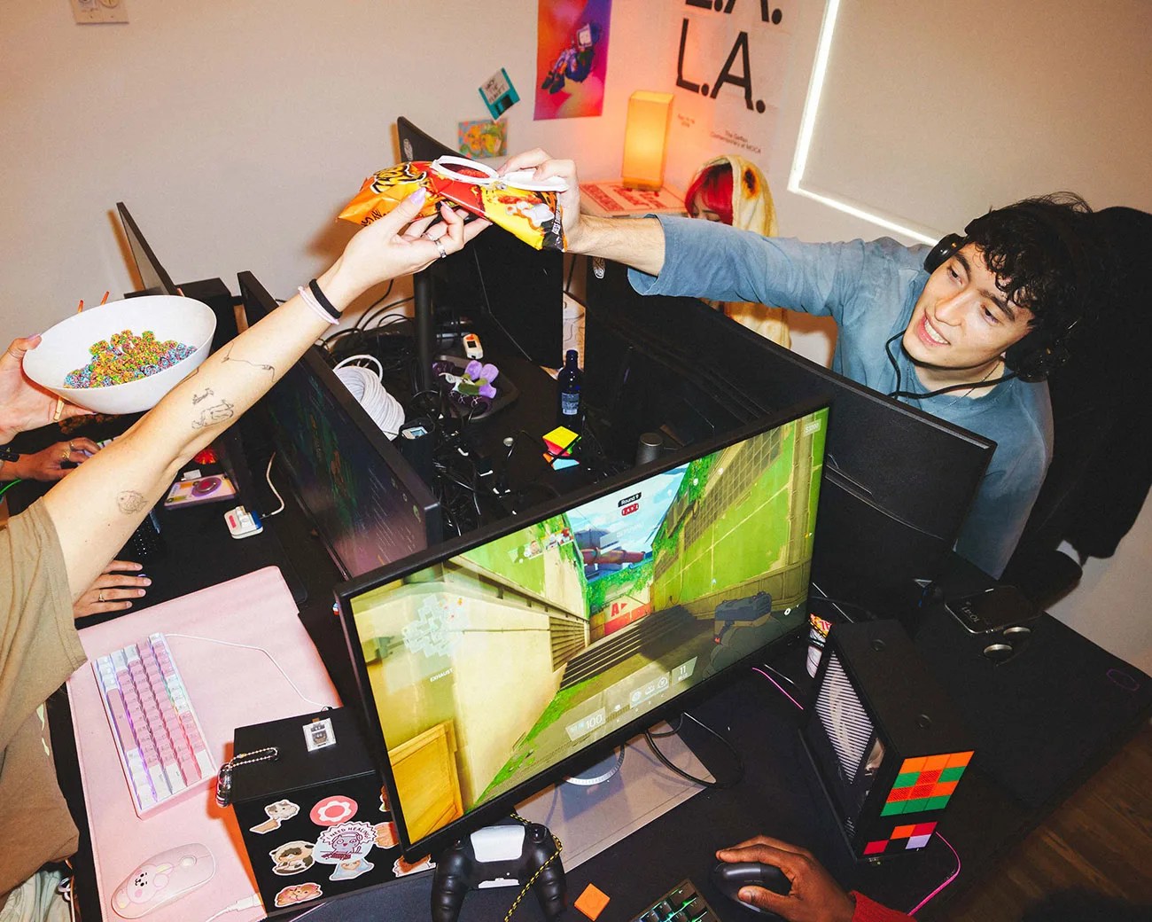 Four people at a gaming setup with multiple monitors, one holding a bowl of colorful cereal and another handing over a bag of chips.