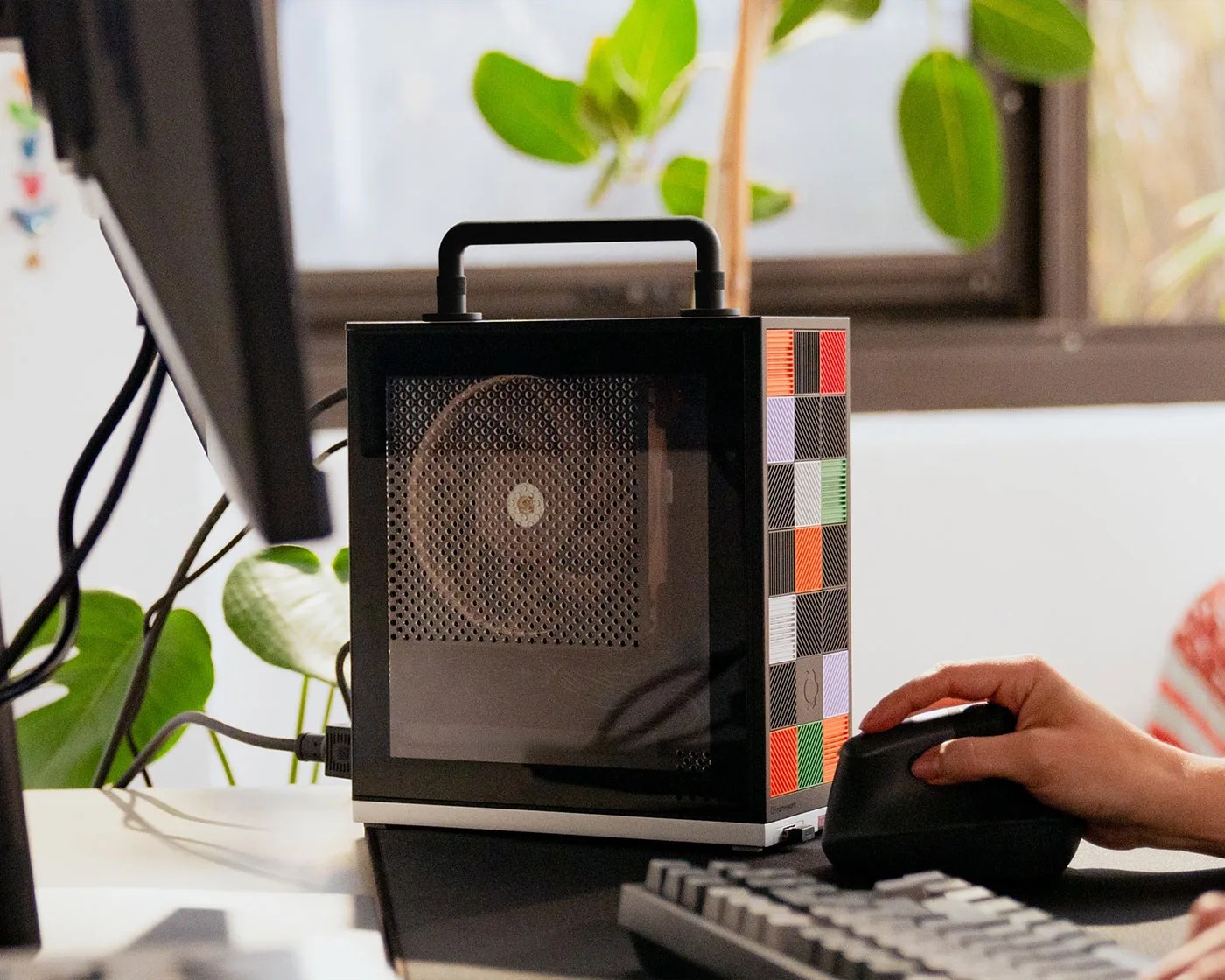 Compact black computer case with a handle and colorful square patterns on the side, placed on a desk next to a keyboard and a hand using a vertical mouse.