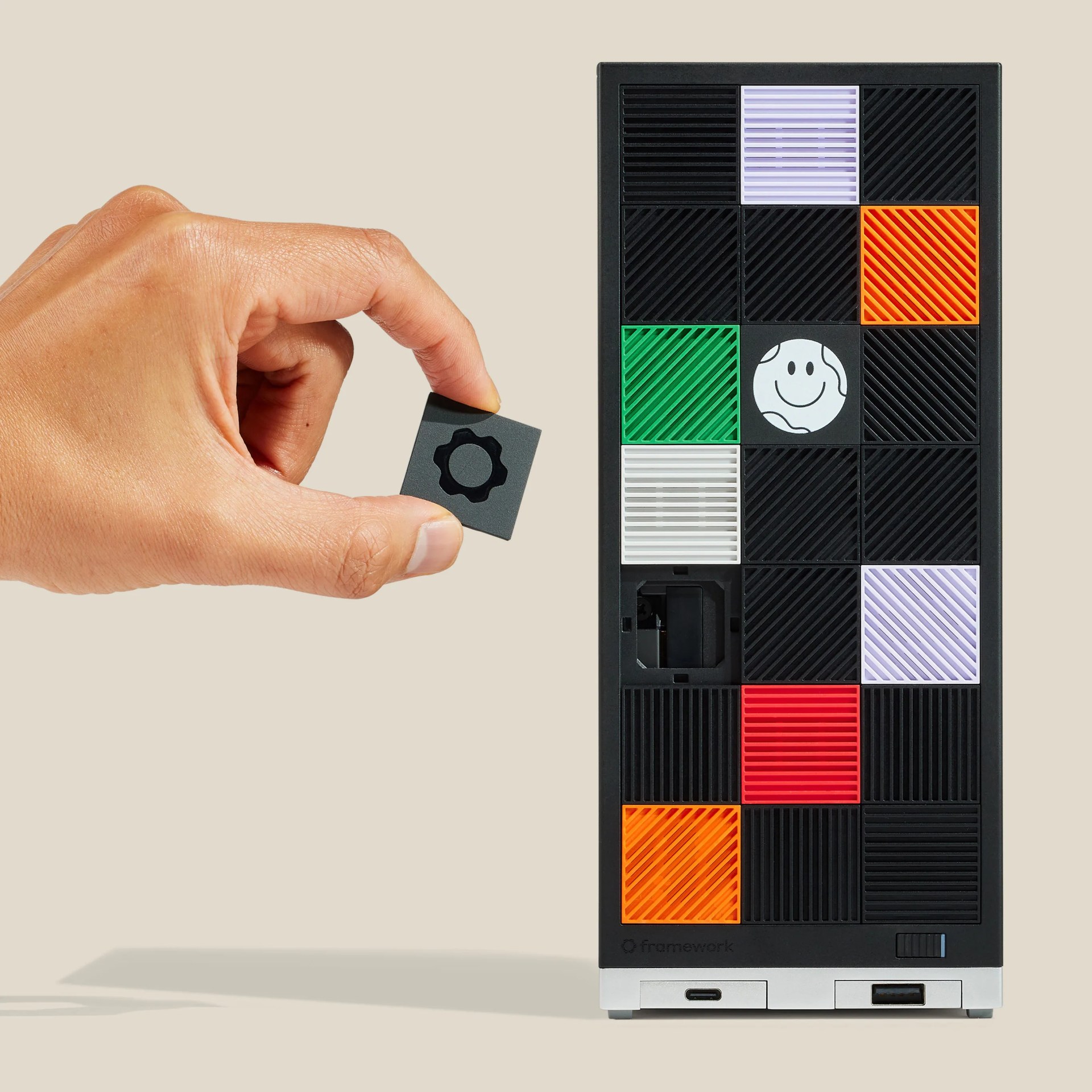 Hand holding a small black square module with a gear icon next to a black rectangular device with colorful modular tiles.