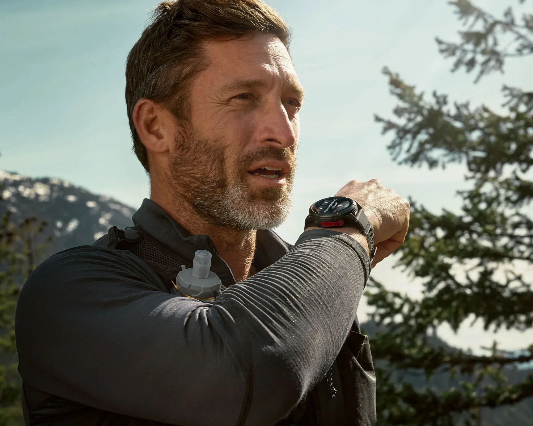 Man wearing a black smartwatch with a red button on his wrist outdoors near trees and mountains.