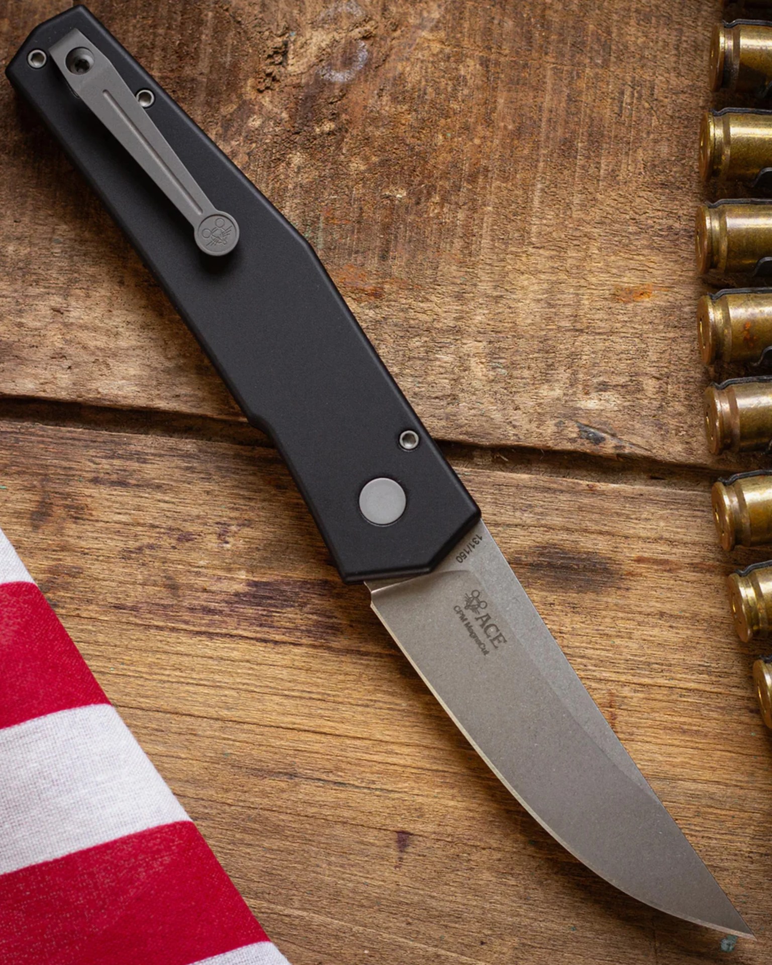 Folding knife with black handle and stonewashed blade on a wooden surface next to a red and white striped cloth and brass bullet casings.