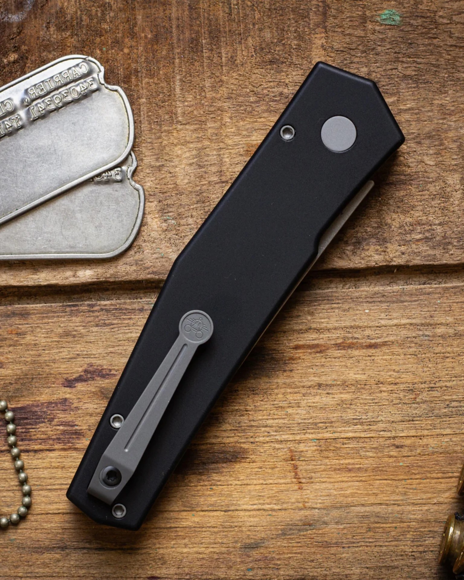 Black folding pocket knife with a gray metal clip on a wooden surface next to metal dog tags.