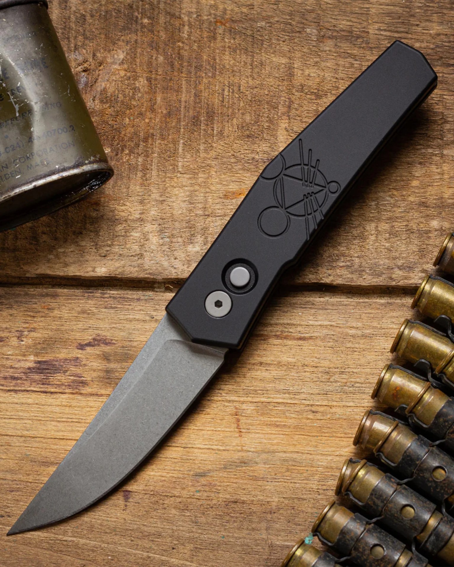 Black folding knife with a stonewashed blade on a wooden surface next to a belt of brass bullets.