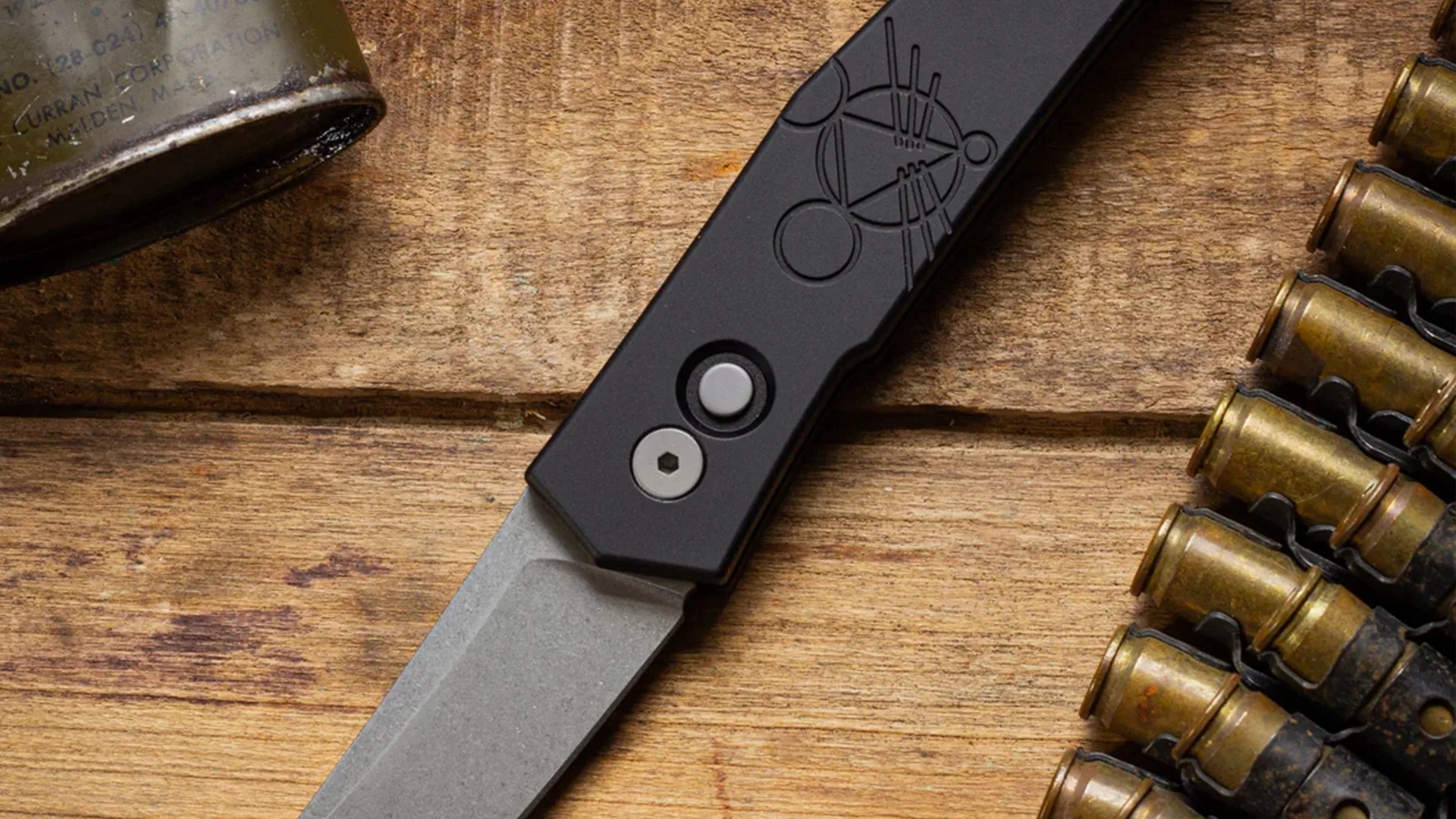 Black folding knife with a stonewashed blade and geometric design on the handle, on a wooden surface next to brass bullet casings.