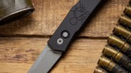 Black folding knife with a stonewashed blade and geometric design on the handle, on a wooden surface next to brass bullet casings.