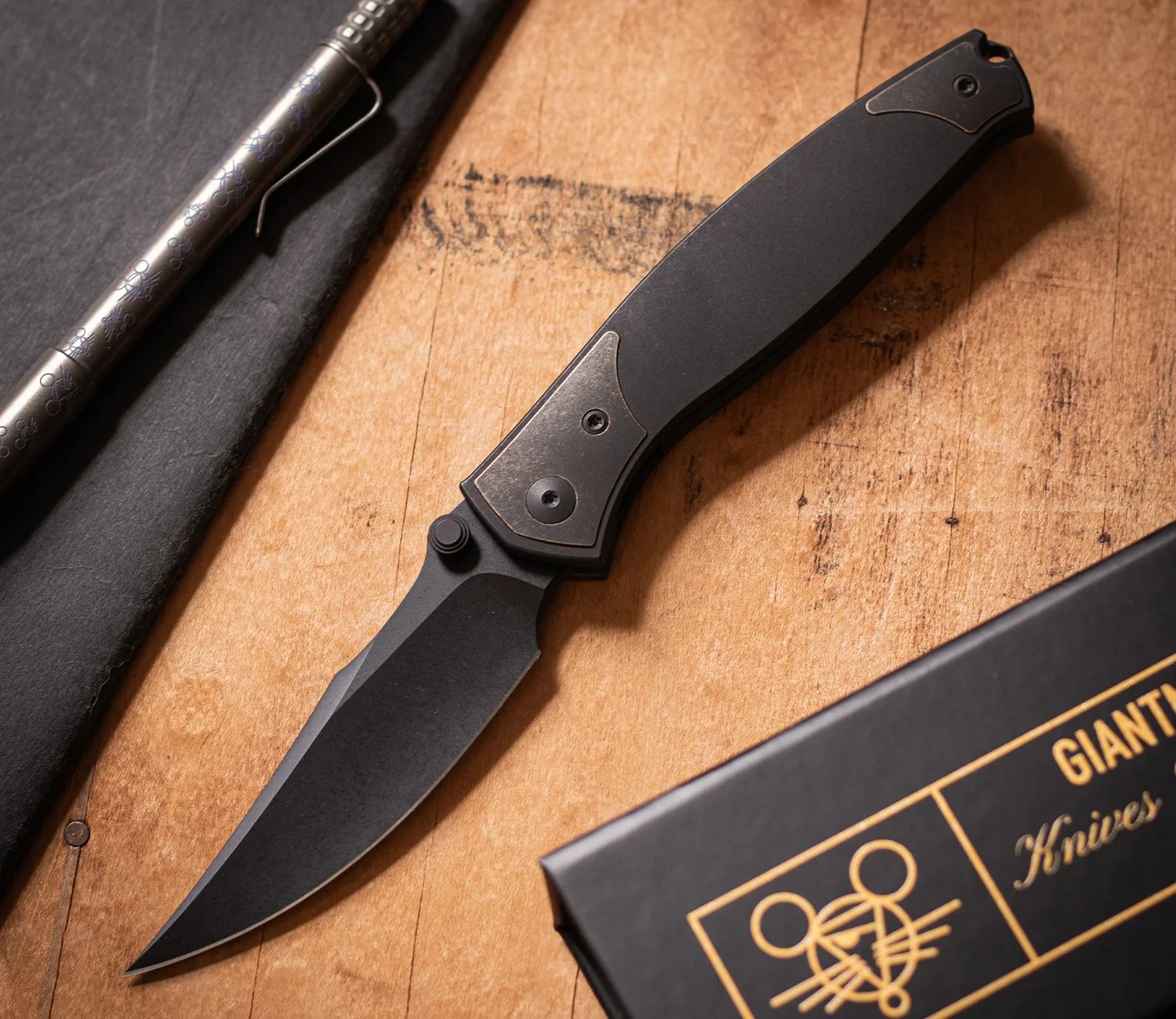 Black folding knife with a matte blade and textured handle on a wooden surface next to a black box and silver pen.
