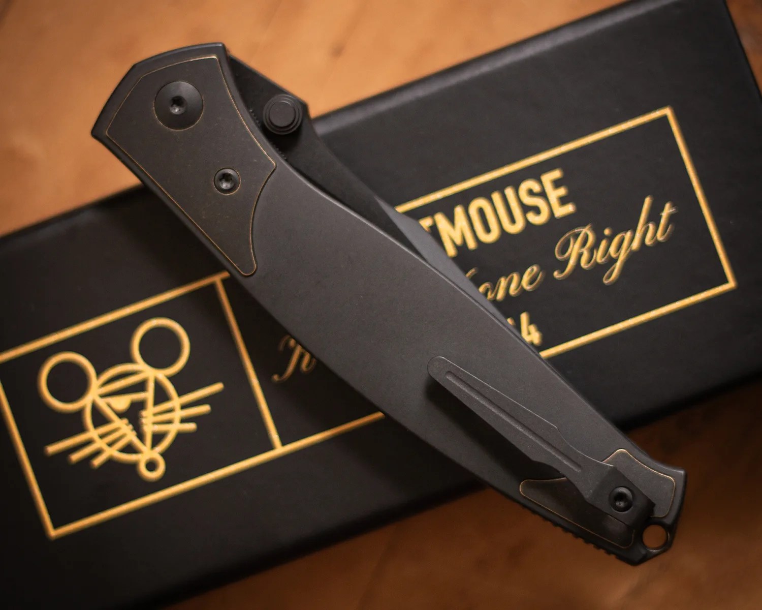 Closed black folding knife with a pocket clip resting on a black box with gold mouse logo and text.