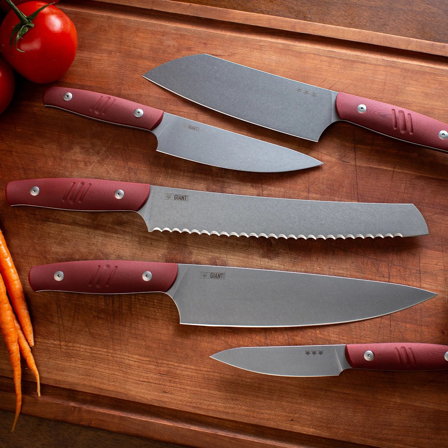 Set of four kitchen knives with gray blades and red handles on a wooden cutting board with tomatoes and carrots.