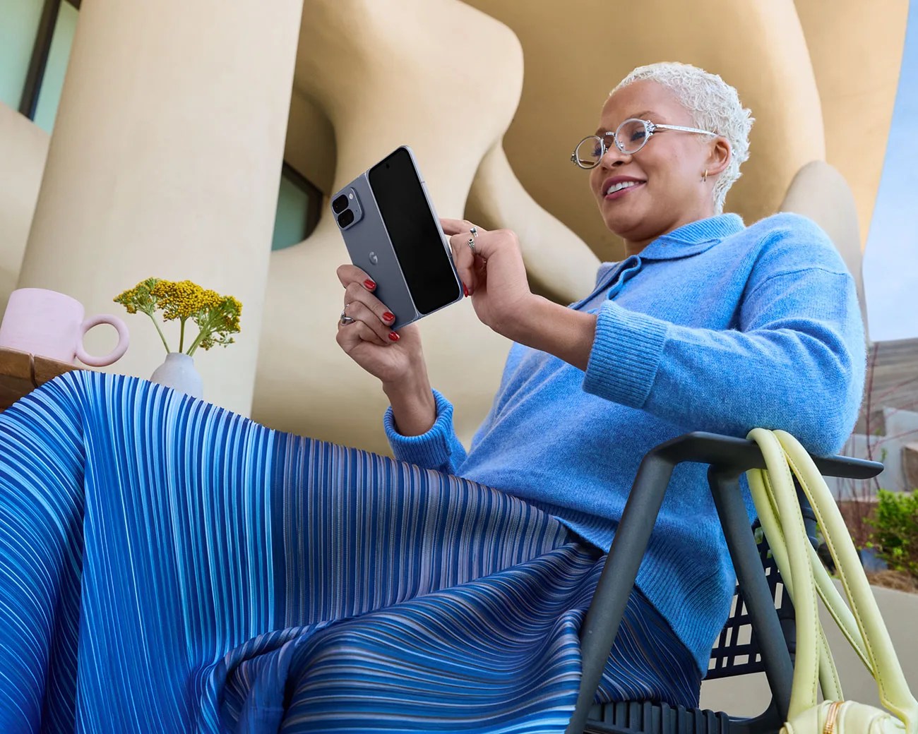 Person in blue sweater and striped blue skirt sitting on a chair, holding a gray foldable smartphone.