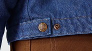 Close-up of blue denim jacket hem with copper button labeled "Carhartt's" over brown textured fabric.