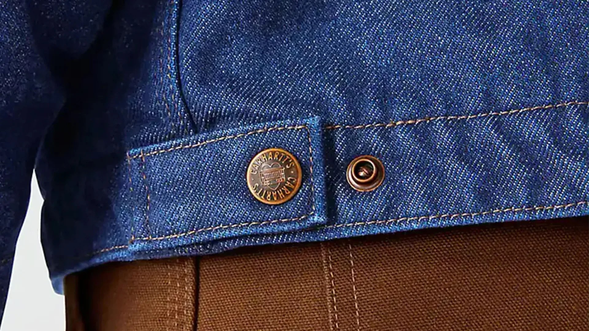 Close-up of blue denim jacket hem with copper button labeled "Carhartt's" over brown textured fabric.