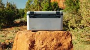 Light gray Dometic cooler with black latches on a large orange rock in a desert landscape.