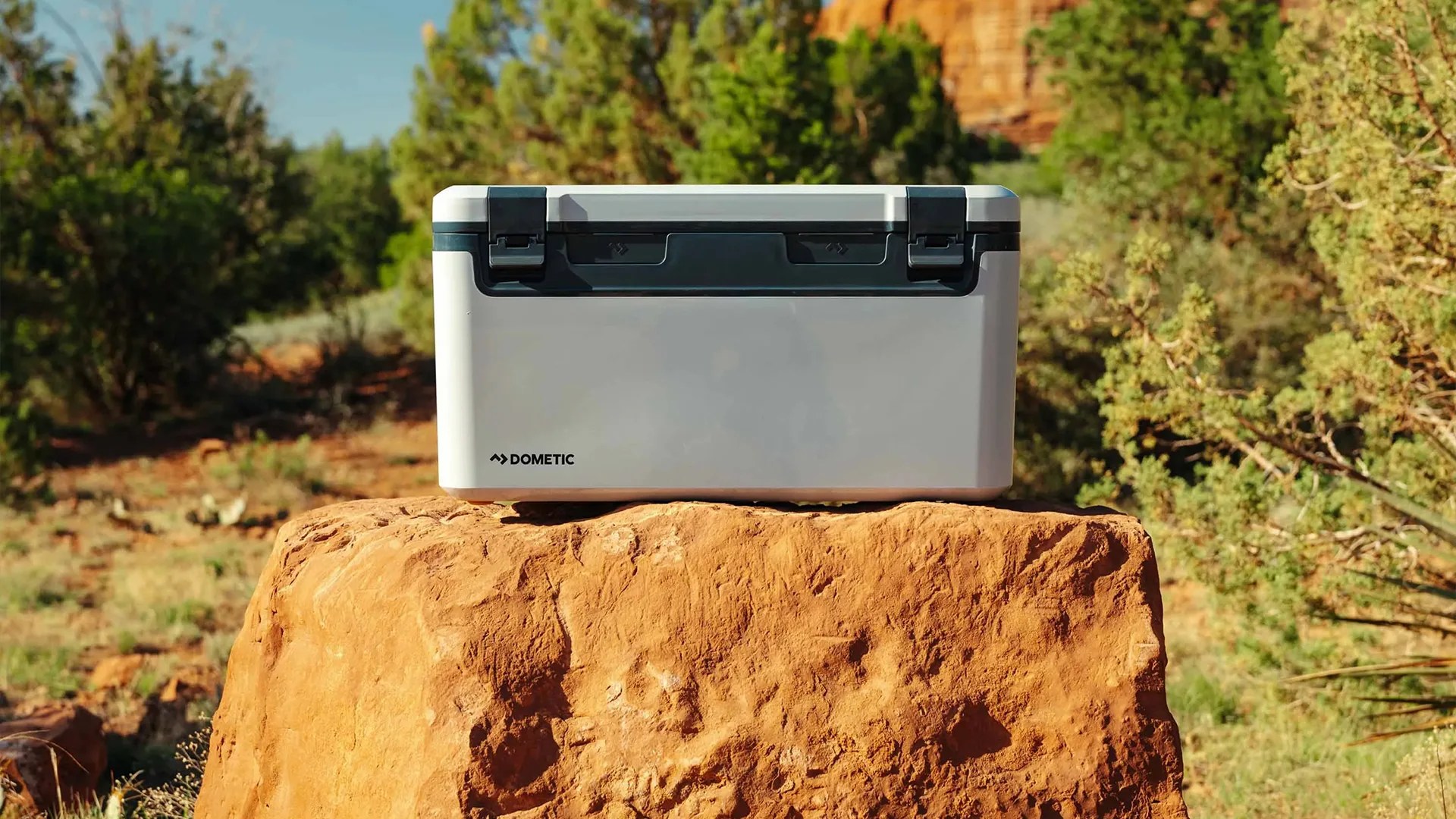 Light gray Dometic cooler with black latches on a large orange rock in a desert landscape.