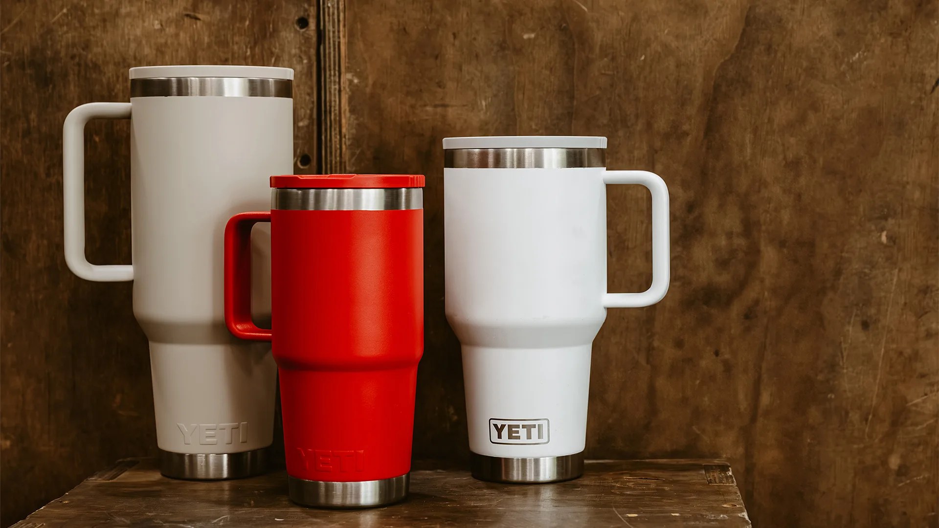 Three YETI insulated mugs in white, red, and light gray with handles, placed on a wooden surface against a brown wooden background.
