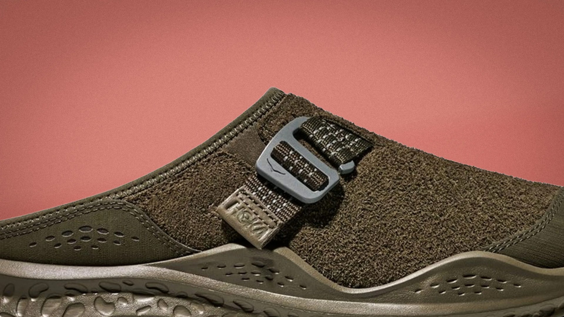 Olive green slip-on shoe with textured upper and adjustable strap buckle on a pink background.
