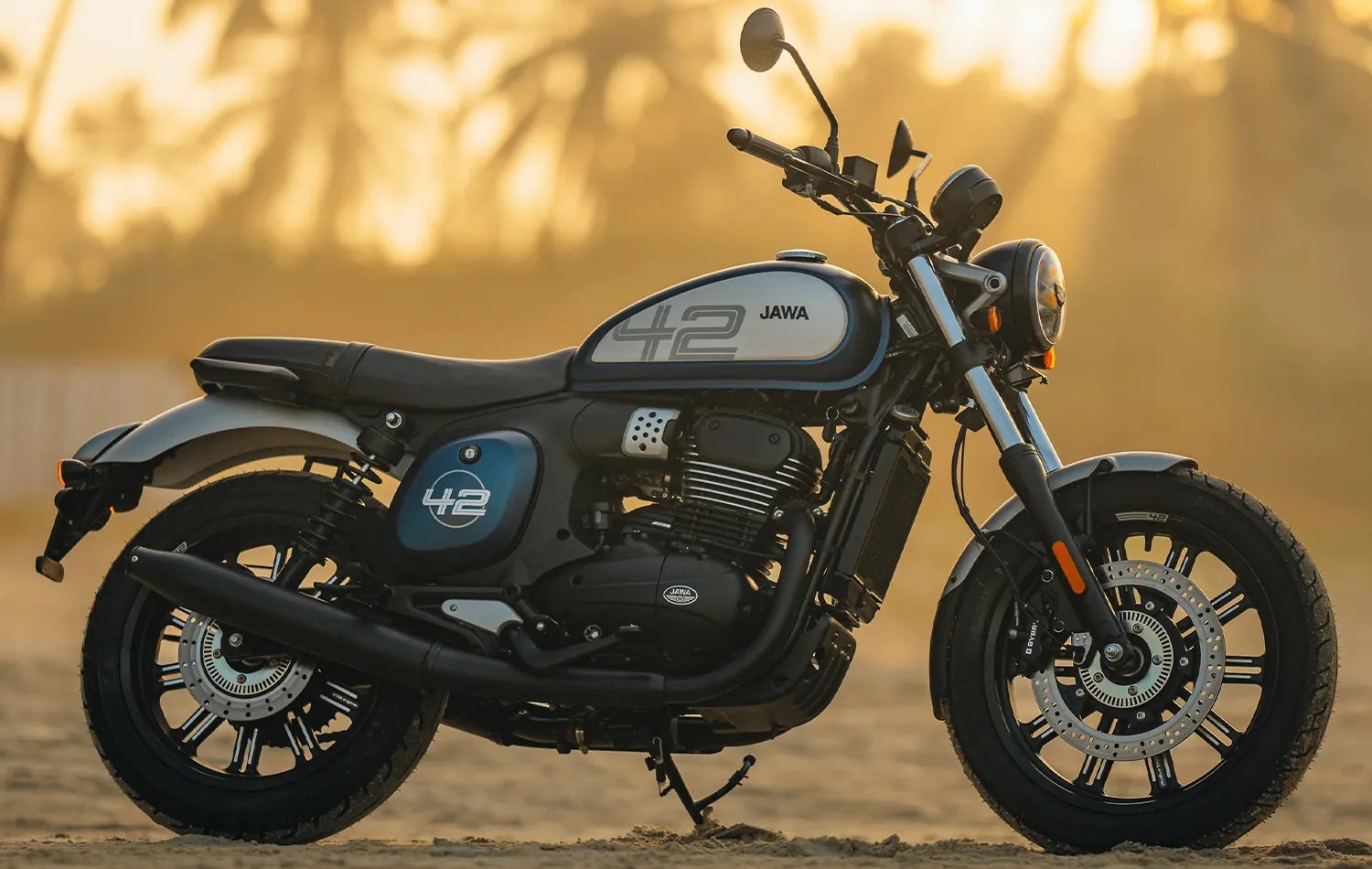 Black and silver Jawa 42 motorcycle with blue side panel and number 42 on fuel tank, parked on sandy ground.