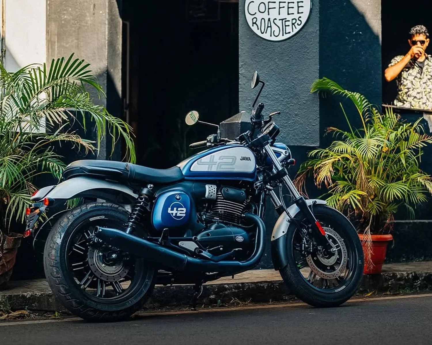 Blue and silver Jawa 42 motorcycle parked on a street near potted plants and a coffee roasters sign.