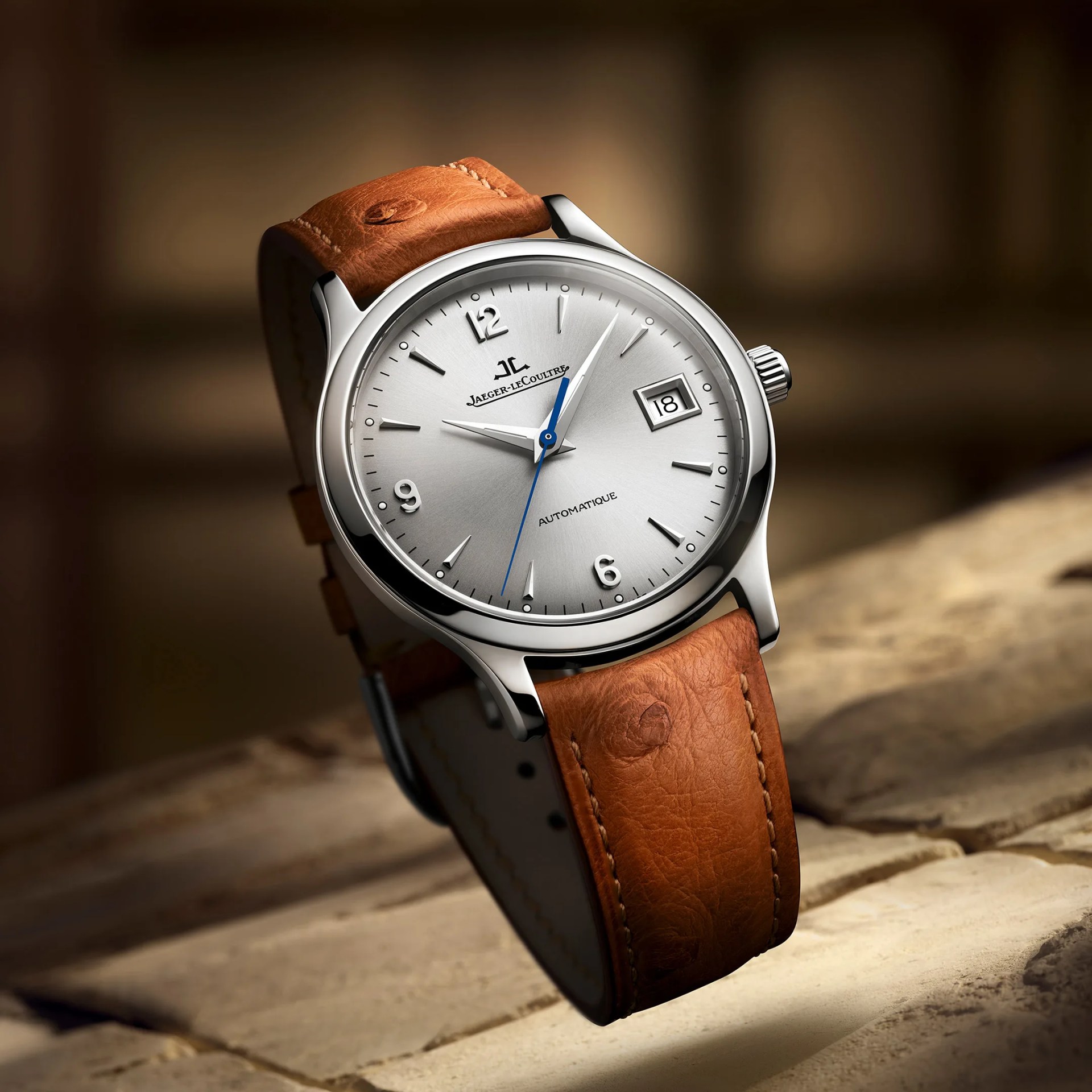 Silver Jaeger-LeCoultre watch with blue second hand and brown leather strap on a stone surface.