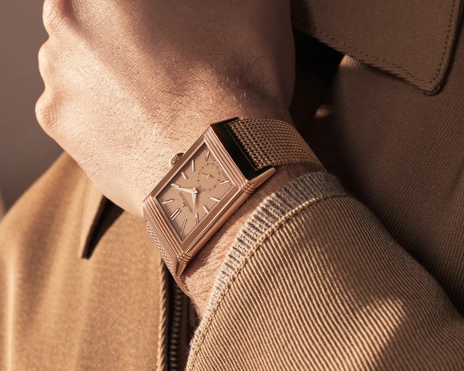 Rectangular rose gold wristwatch with mesh strap worn on a wrist over a brown jacket sleeve.