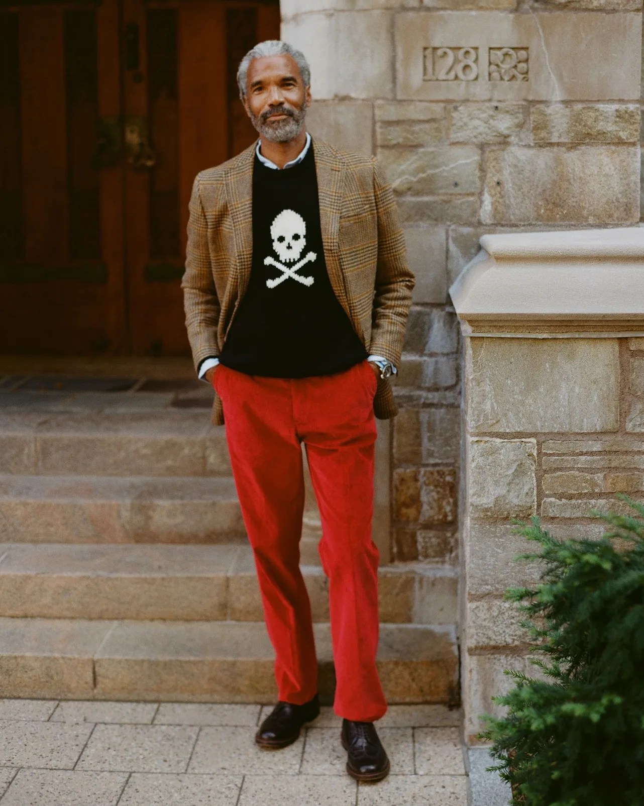 Man wearing a brown plaid blazer, black sweater with white skull and crossbones, red pants, and black shoes standing on stone steps.