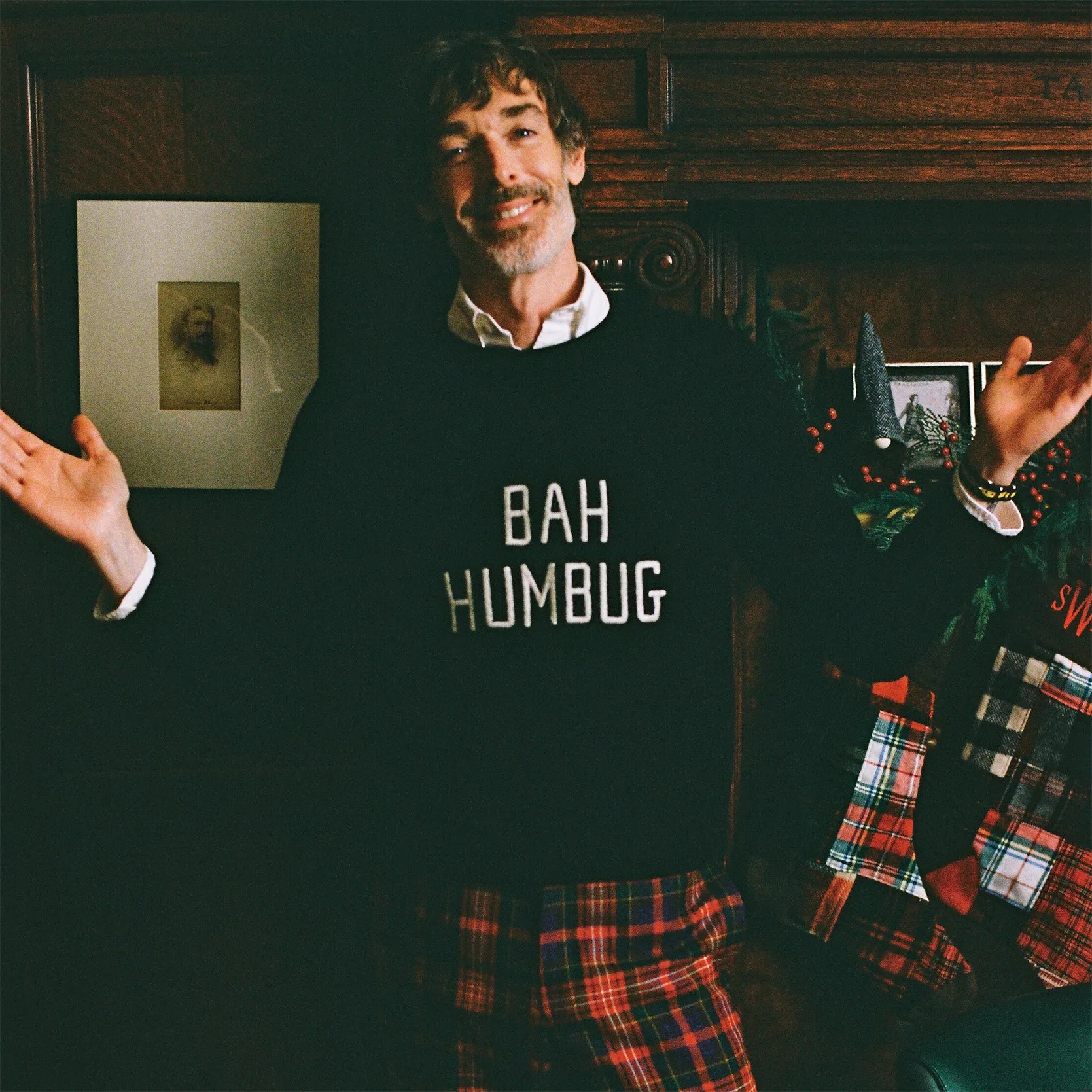 Man wearing a black sweater with "BAH HUMBUG" text and red plaid pants standing with arms open.