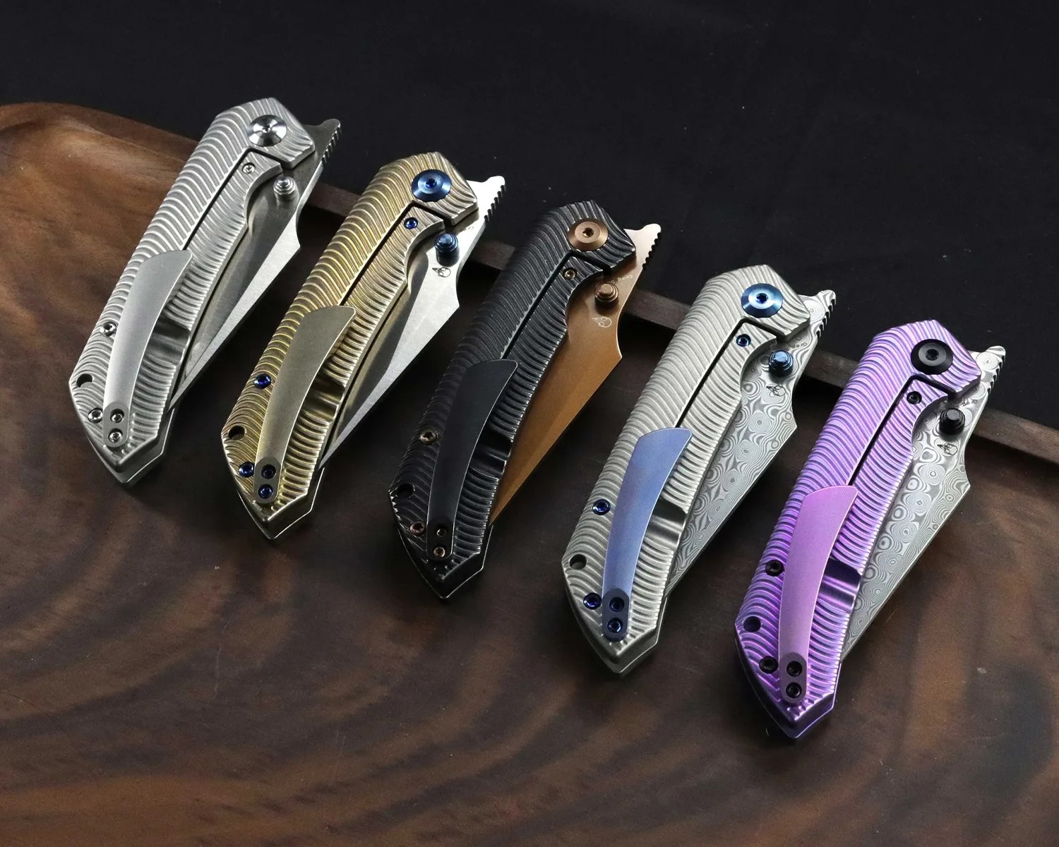 Five closed folding knives with textured handles in silver, gold, black, silver with blue clip, and purple, arranged on a wooden surface.