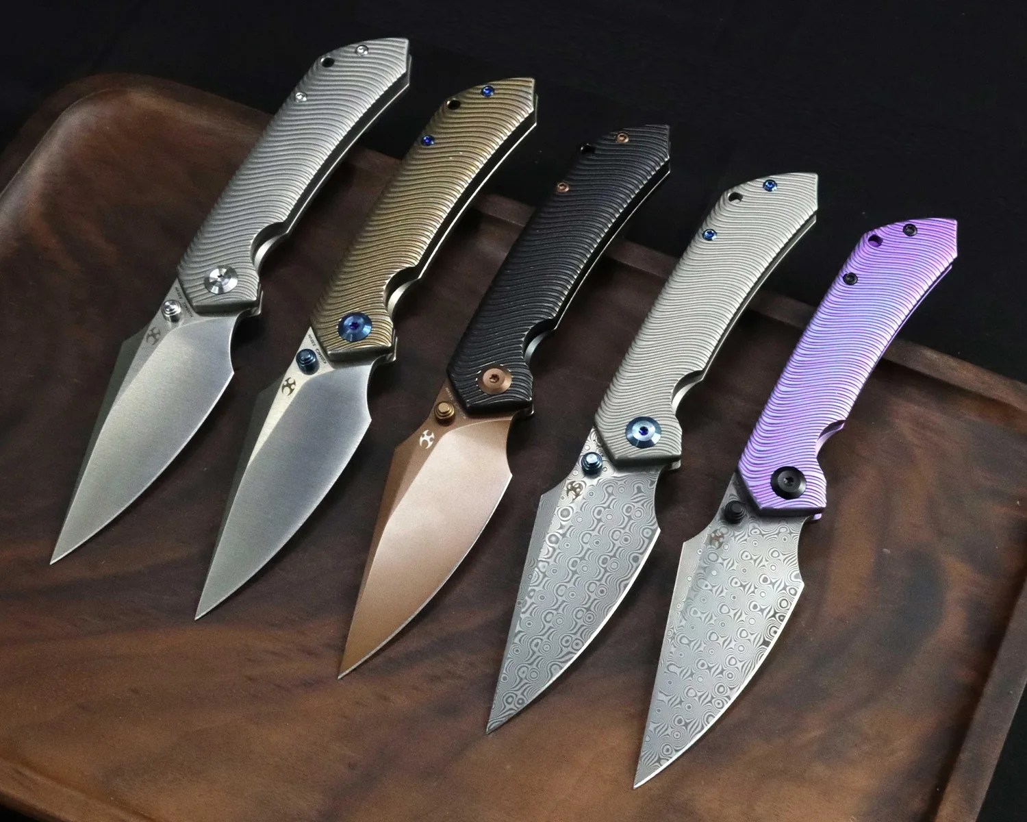 Five folding knives with textured handles in silver, gold, black, silver, and purple, displayed on a wooden tray.