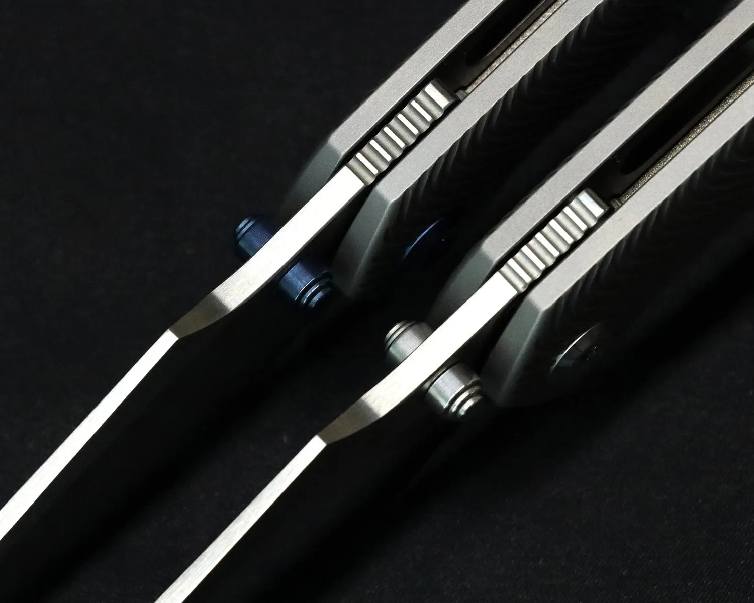 Close-up of two folding knives showing the spine, thumb studs (one blue, one silver), and textured metal handles on a black background.