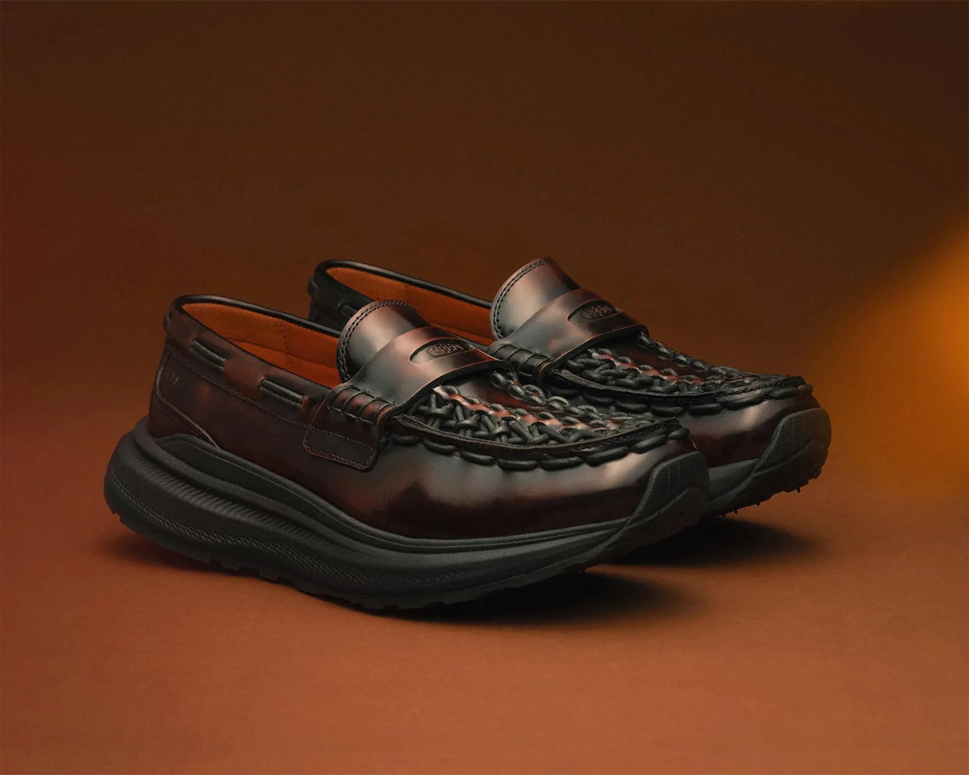 Pair of dark brown leather woven moccasin loafers with thick black rubber soles on a brown surface.
