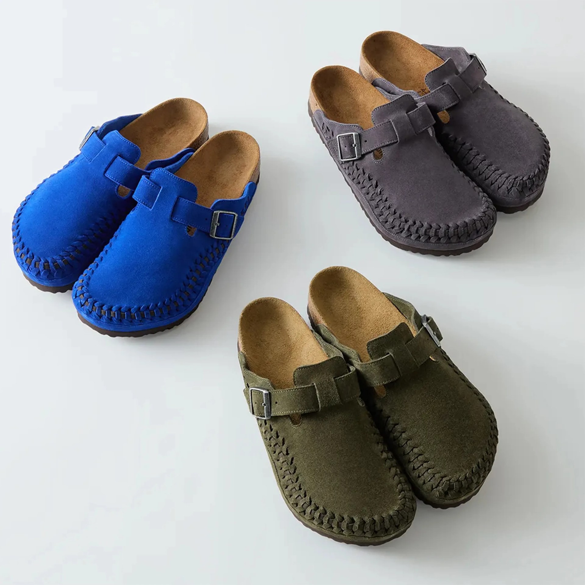 Three pairs of suede slip-on clogs with buckled straps in blue, gray, and olive green on a white surface.