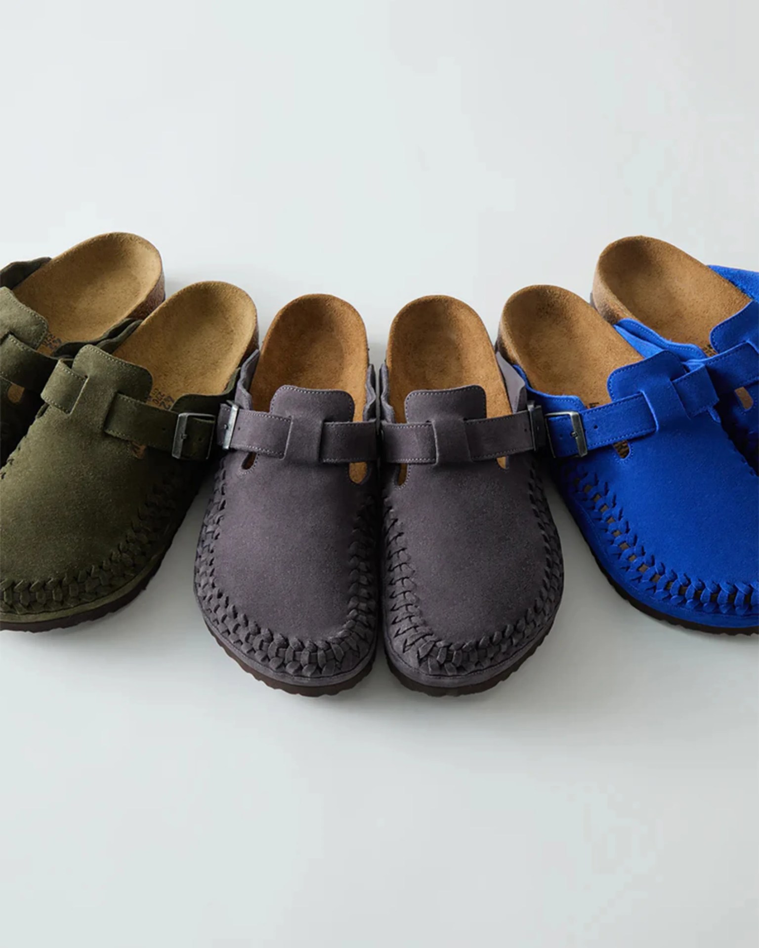 Three pairs of suede slip-on clogs in olive green, dark gray, and bright blue with woven detailing and buckle straps.