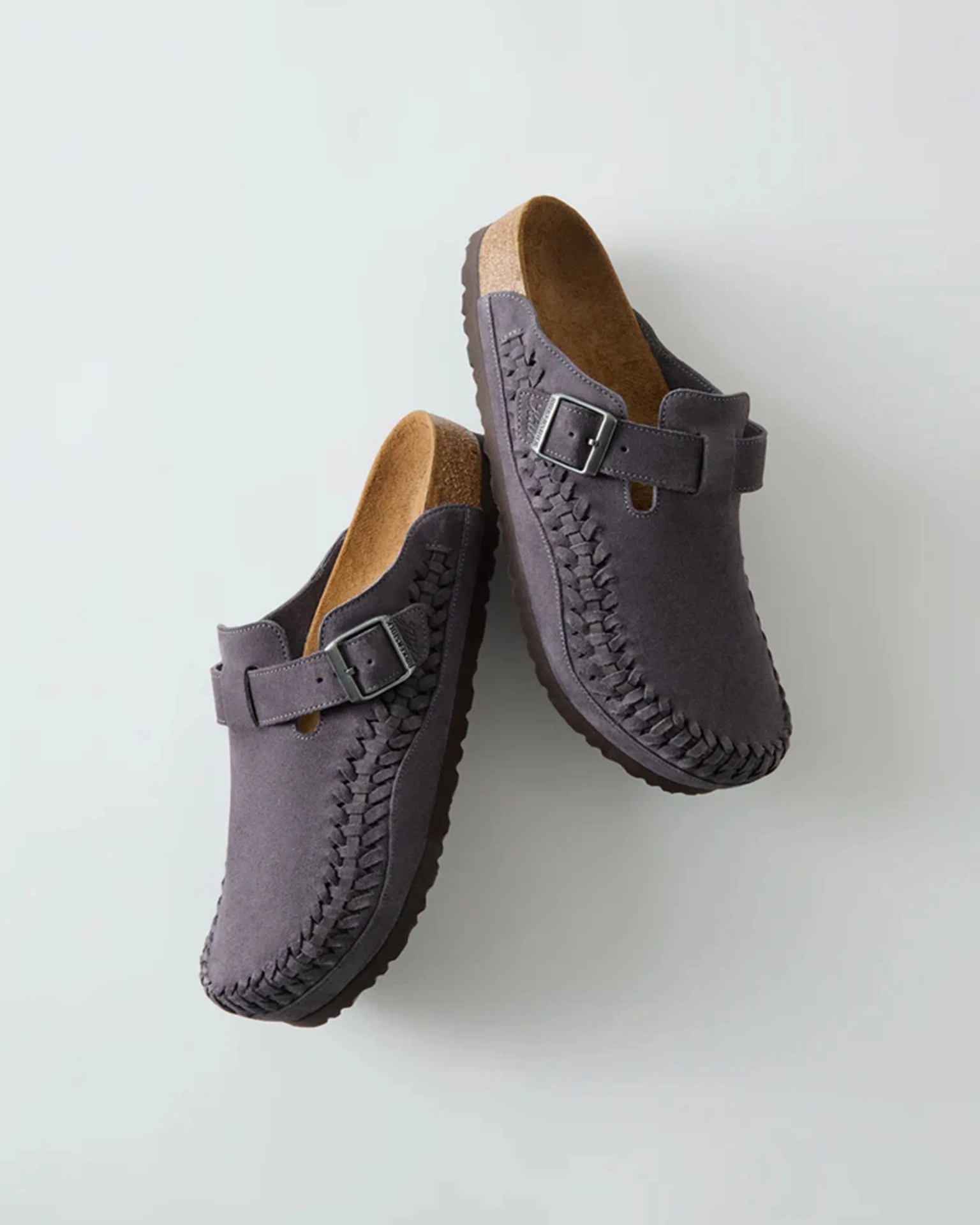 Pair of dark gray suede slip-on clogs with woven detailing and adjustable buckle straps.