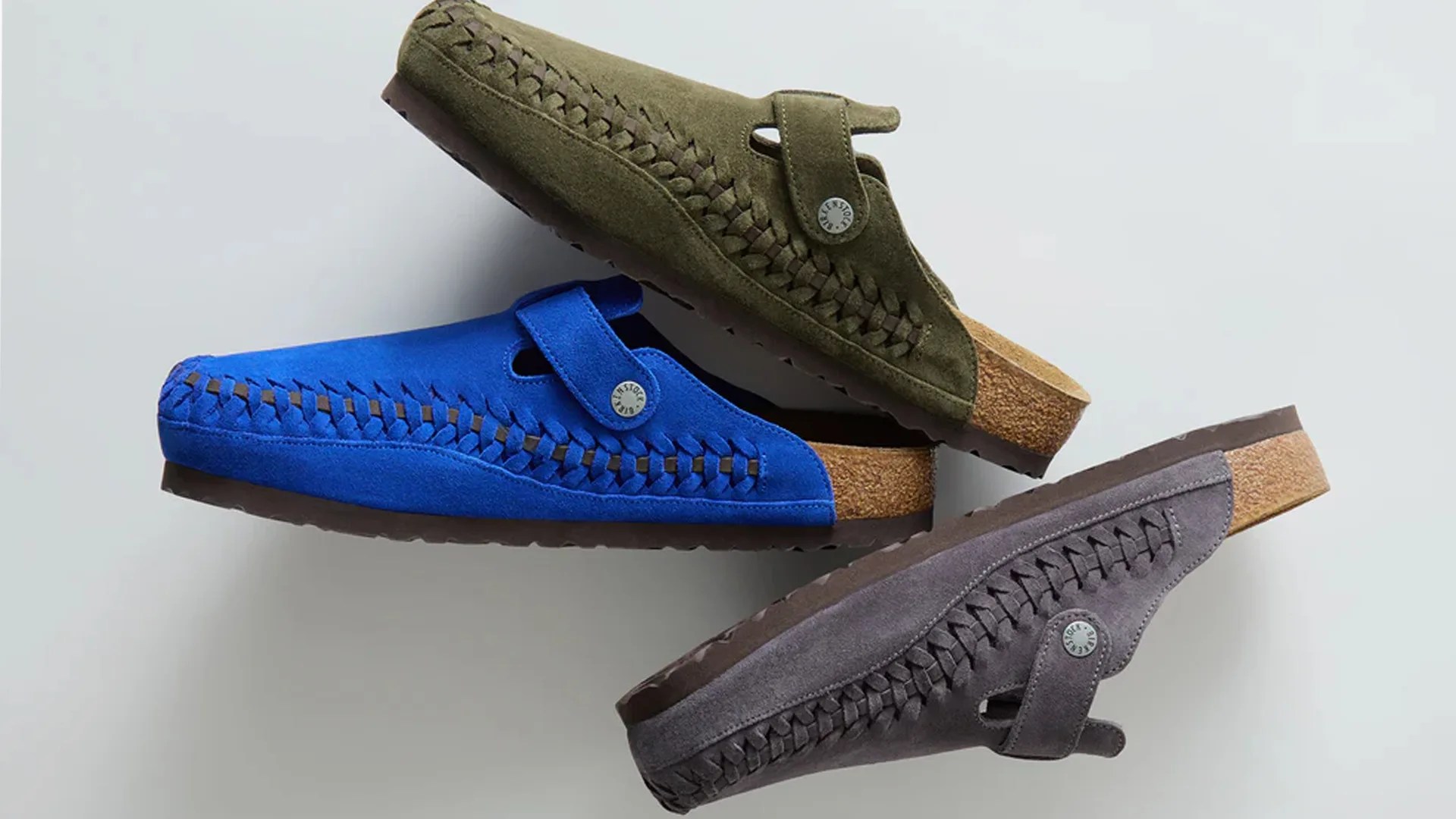 Three suede slip-on clogs in olive green, royal blue, and gray with woven side details and cork soles.
