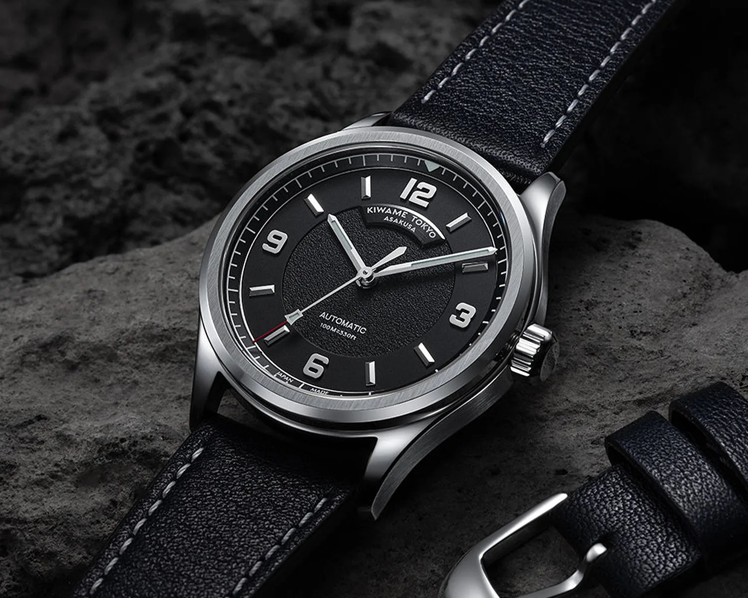 Black dial automatic wristwatch with silver case and black leather strap on a textured stone surface.