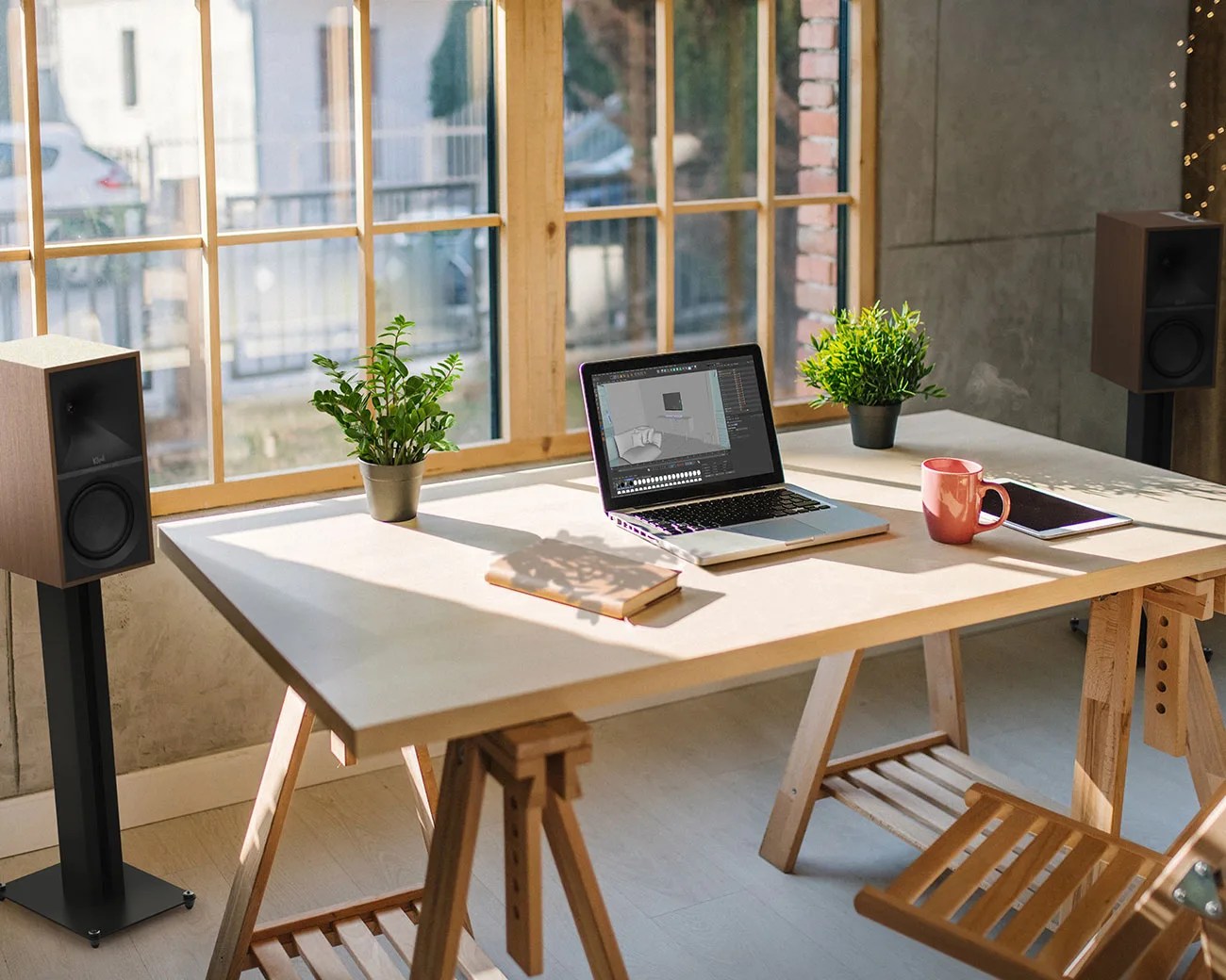 Light wooden desk with a laptop, two potted plants, a pink mug, a notebook, and a tablet near large windows.