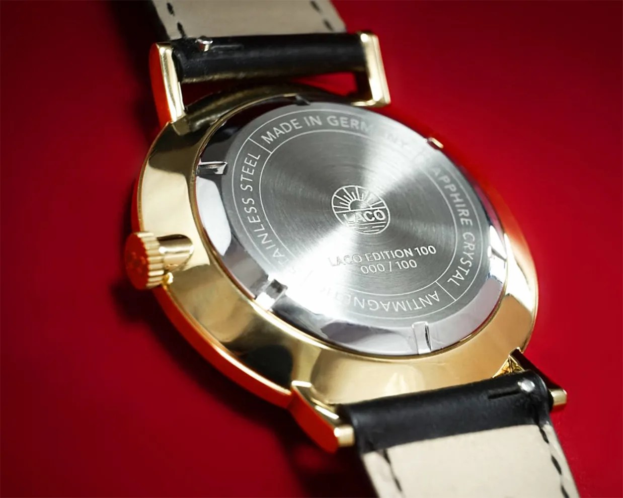 Close-up of the back of a gold watch case with stainless steel cover, black leather strap, and engraved text on a red background.