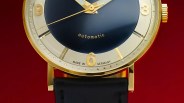 Close-up of a gold watch with a black dial, gold numerals, and a black leather strap against a red background.