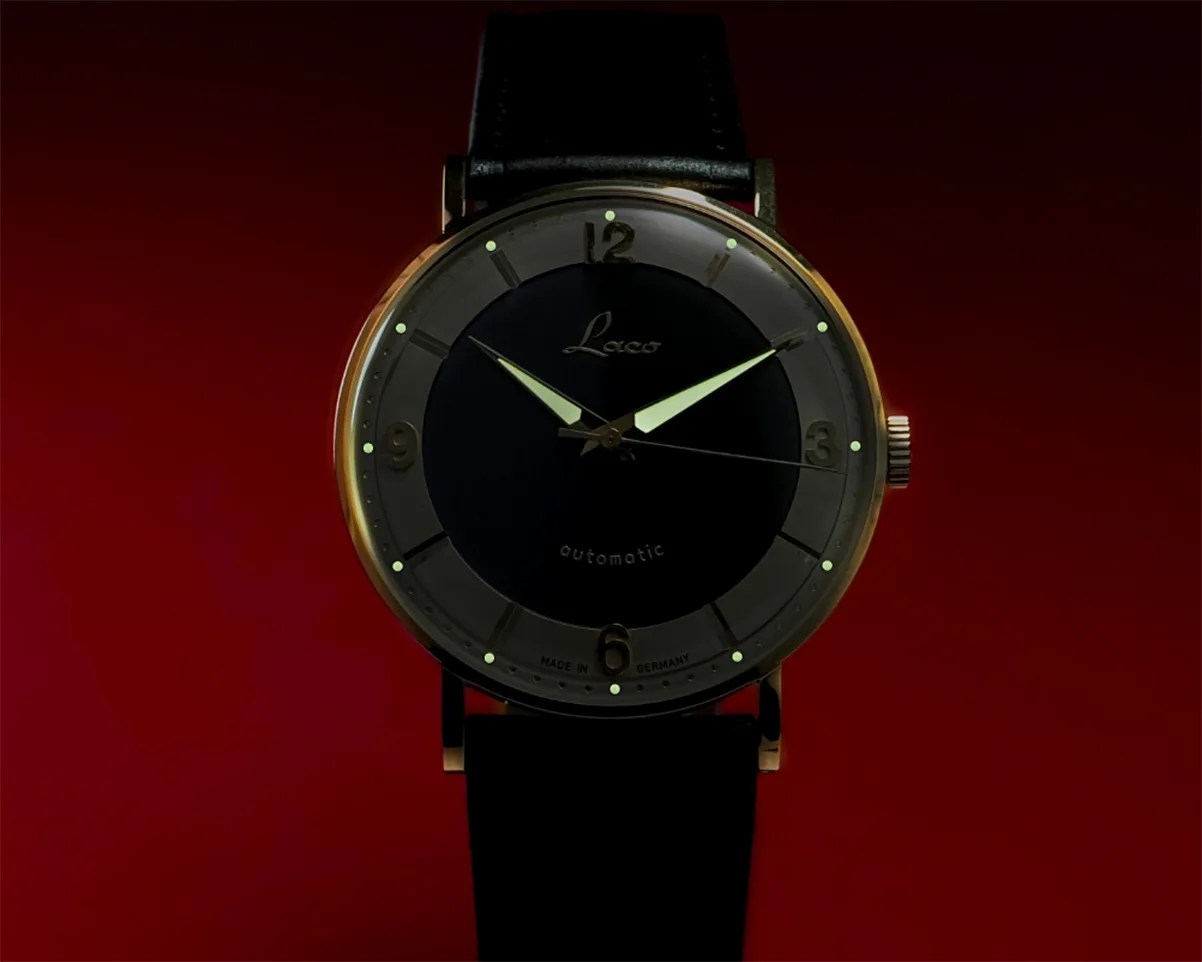 Round wristwatch with black dial, luminous hands, gold-tone case, and black leather strap on a red background.