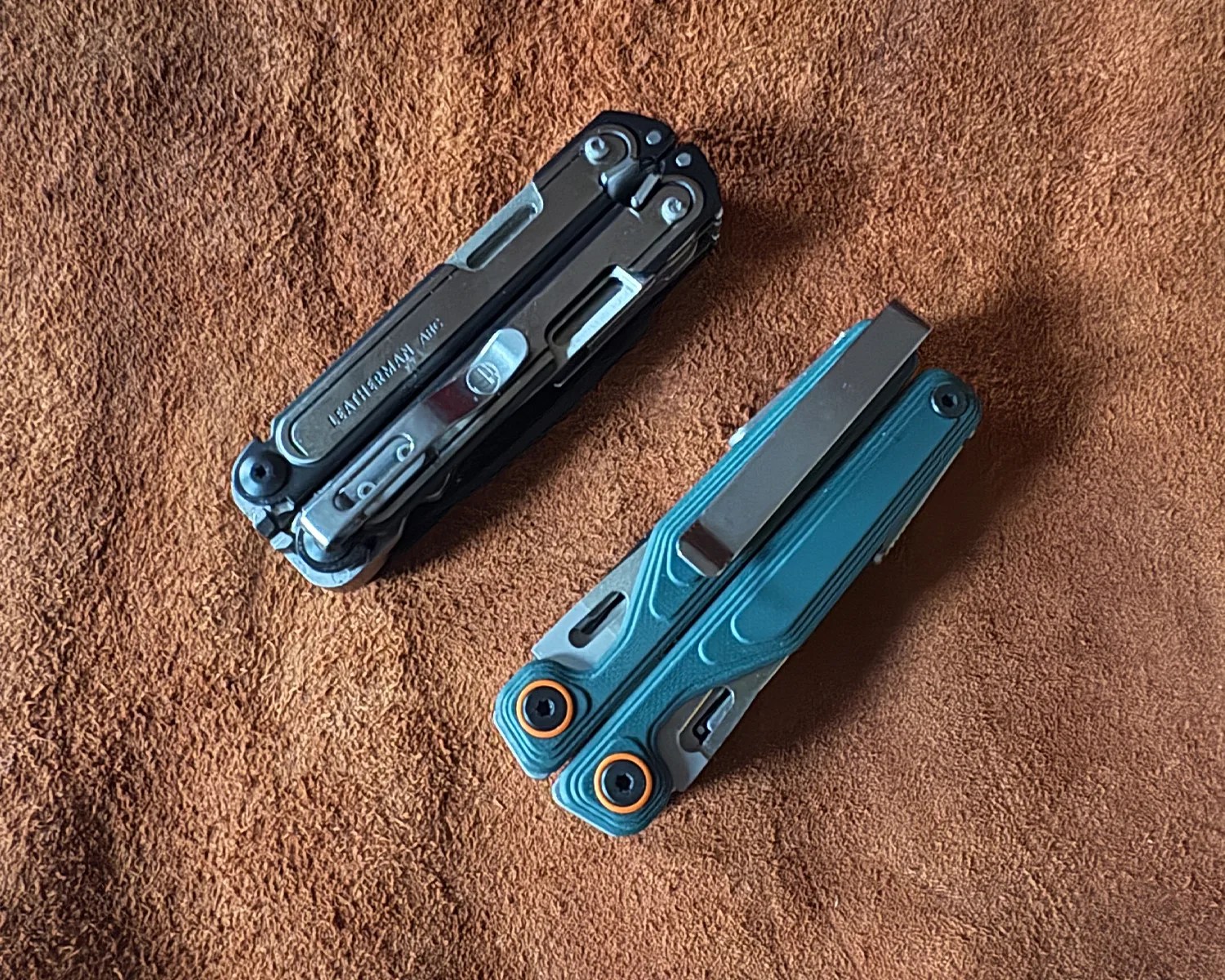 Two folded multitools, one black with "Leatherman" branding and one teal with orange accents, on a brown textured surface.