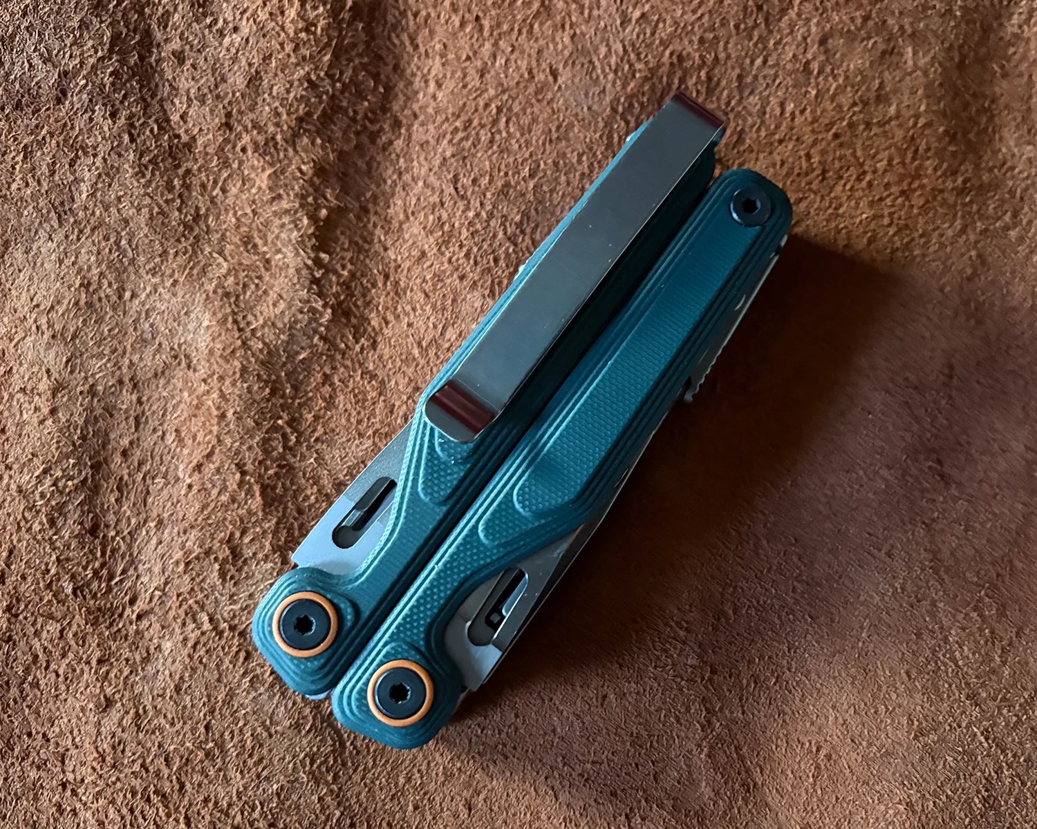 Folded teal multi-tool with black screws and a metal clip on a brown textured surface.