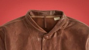 Brown leather jacket with a single button and a stand collar on a red background.