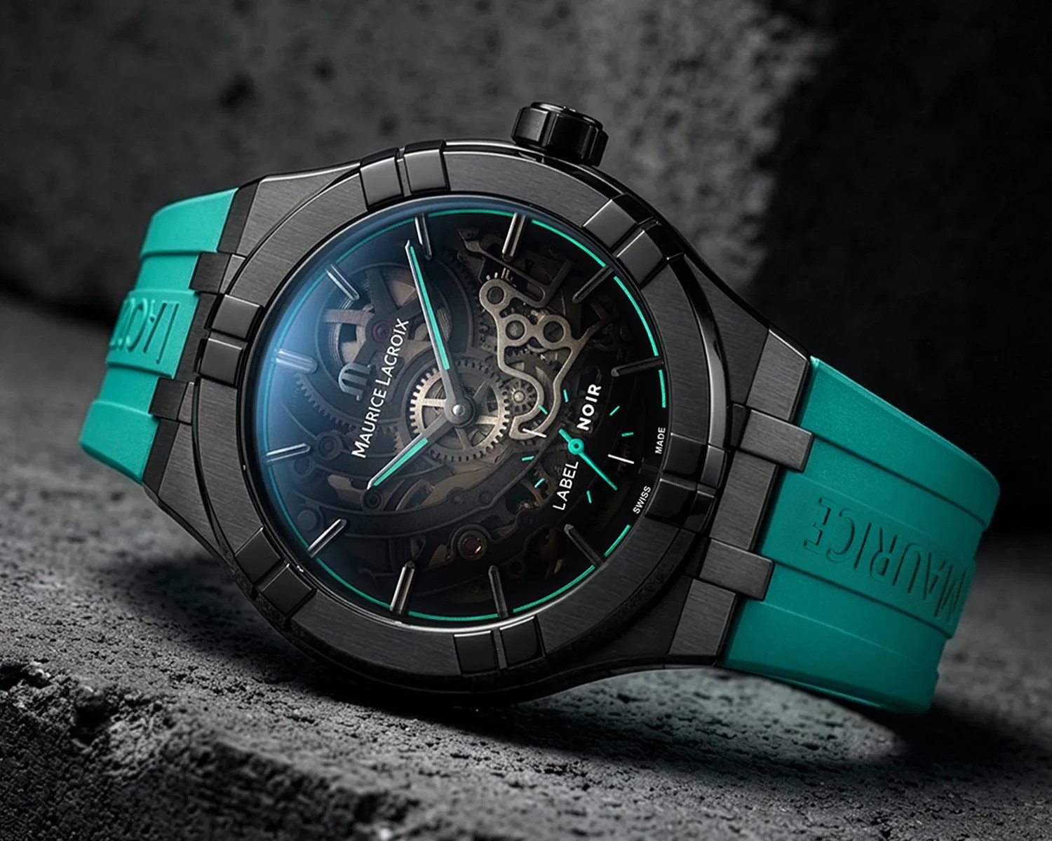 Black Maurice Lacroix wristwatch with visible gears and turquoise rubber strap on textured gray surface.