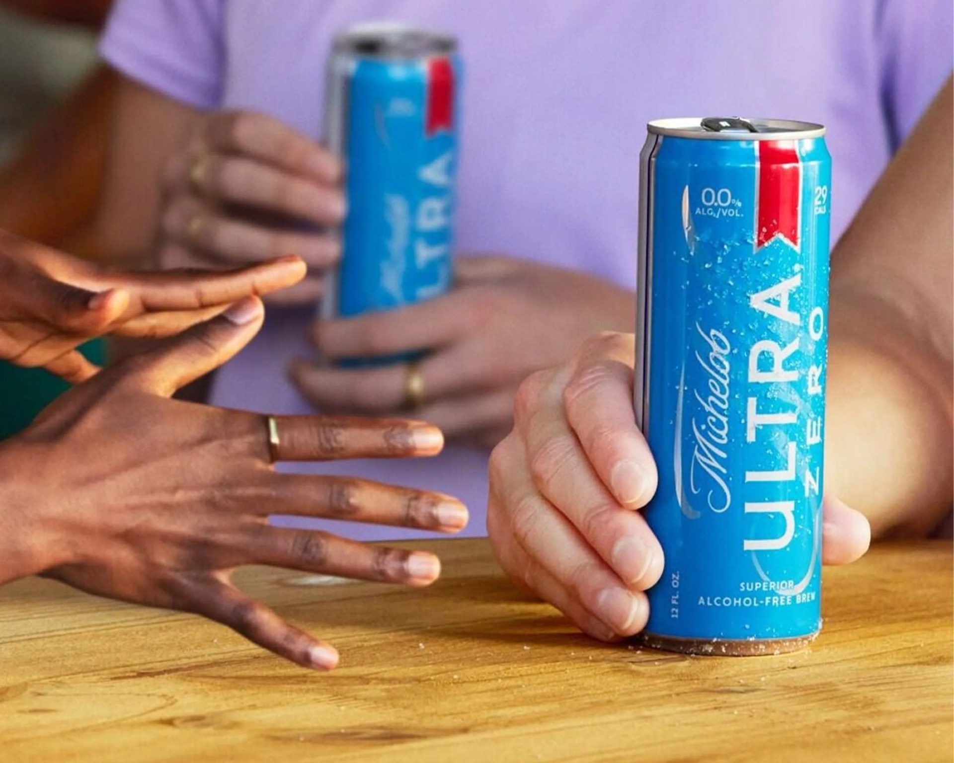 Two hands holding blue Michelob Ultra Zero alcohol-free beer cans over a wooden table.