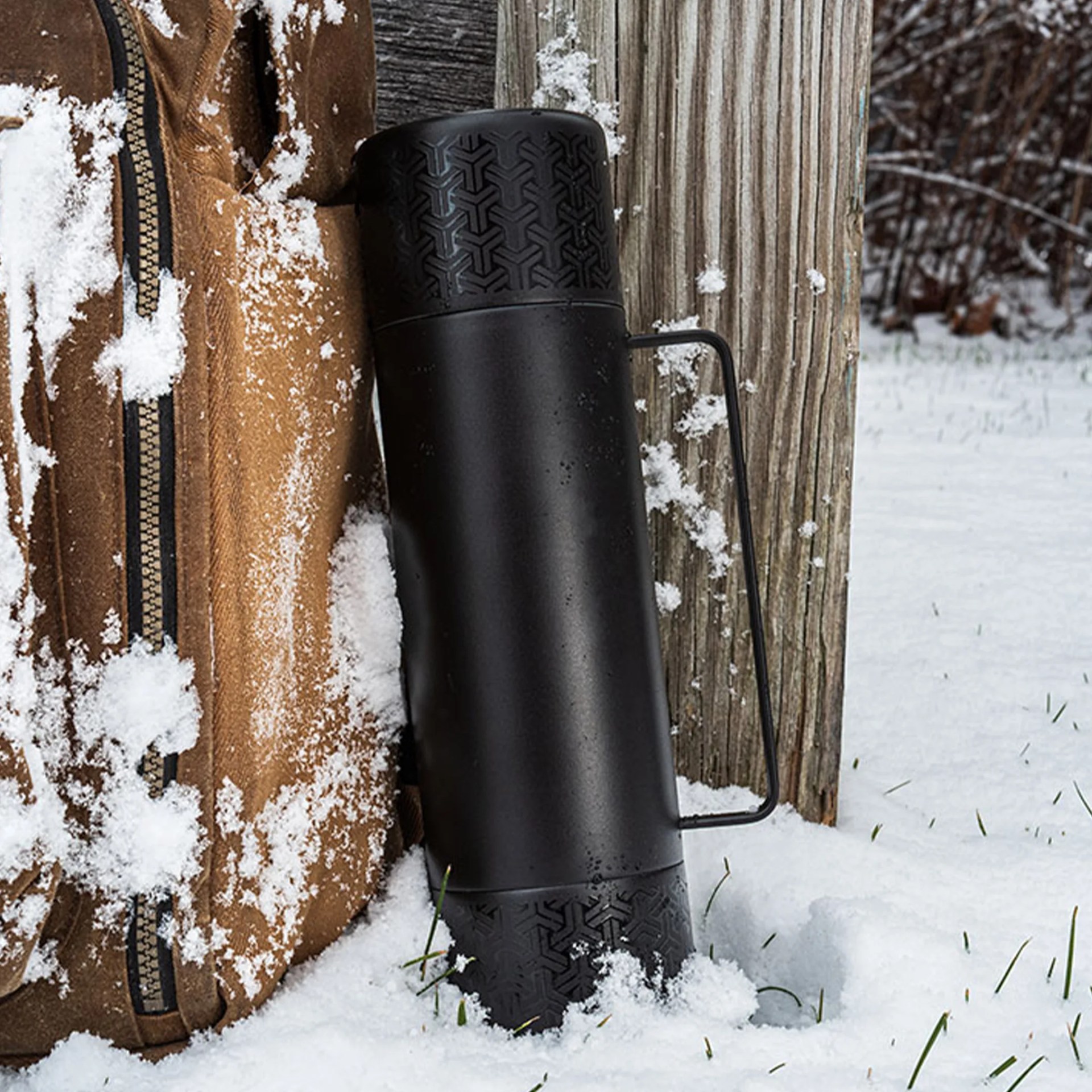 Black metal thermos with textured top and bottom and a handle, standing in snow next to a brown jacket and wooden post.