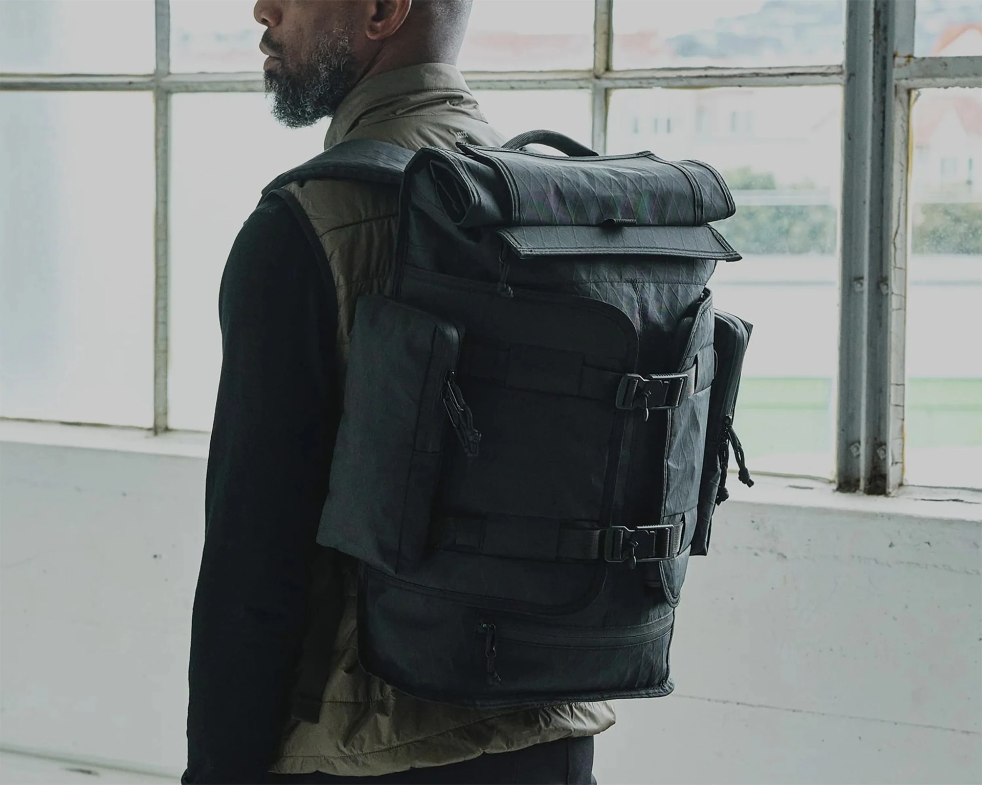 Black roll-top backpack with multiple front pockets and buckles worn by a person standing indoors near large windows.