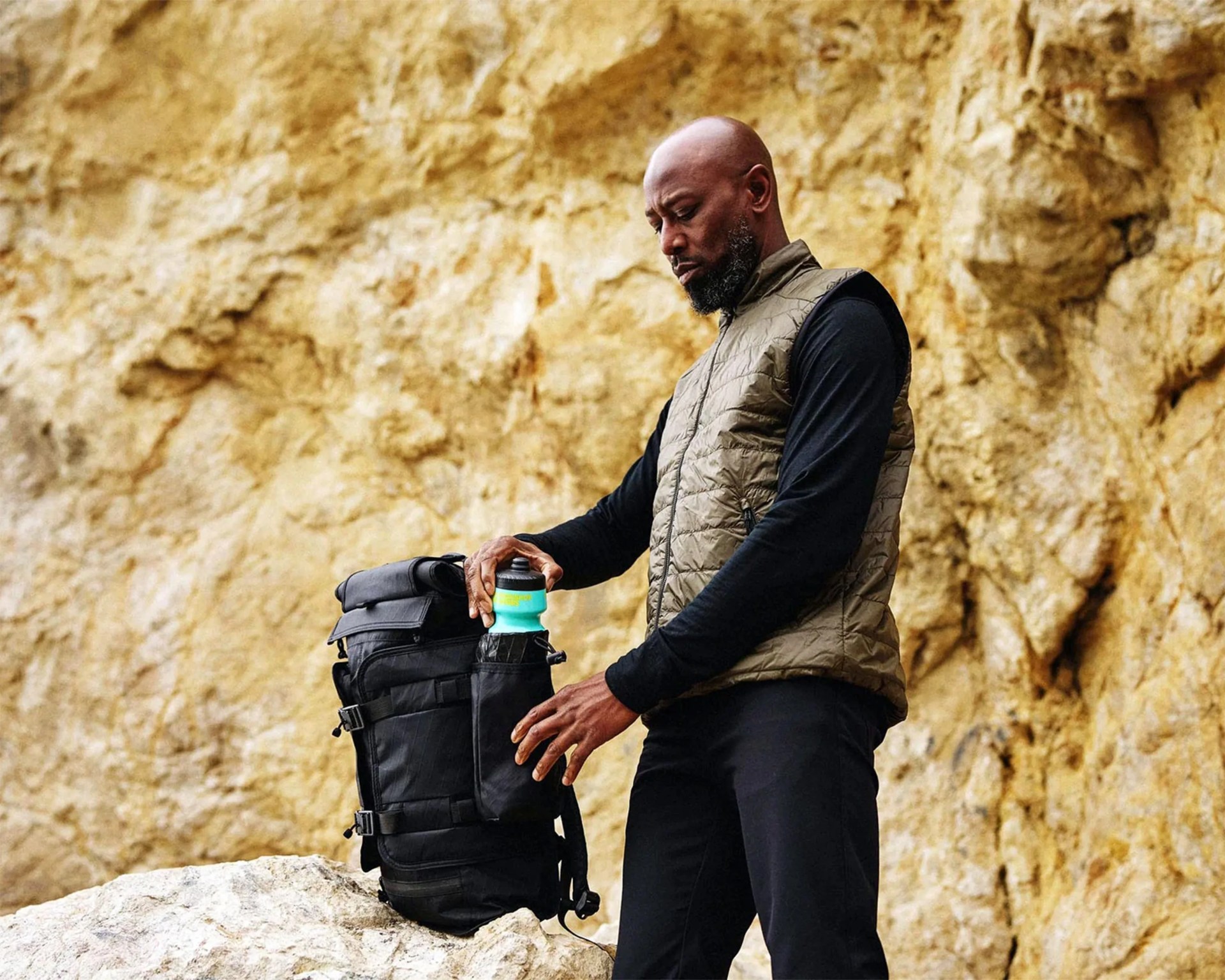 Man placing a turquoise water bottle into the side pocket of a black backpack on a rock with a rocky cliff background.