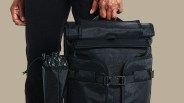 Black roll-top backpack with side pocket holding a folded umbrella, held by a person in black pants.