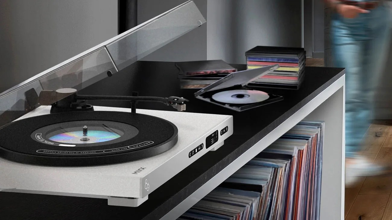White modern turntable with a clear dust cover open, playing a vinyl record, on a black shelf with stacked CDs and vinyl records below.